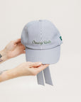 A person holding a blue and white seersucker baseball cap with the text 'Chasing Balls' embroidered on the front and an oversized bow at the back.