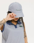 A person wearing a blue and white seersucker baseball cap with the text 'Chasing Balls' embroidered on the front and an oversized bow at the back.