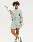 A woman wearing a green striped button-up shirt with a collar, sleeve cuffs, and side seam hem, paired with a matching skirt, sunglasses, and green shoes.