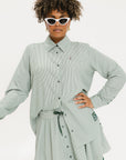 A woman wearing a green striped button-up shirt with a collar, sleeve cuffs, and side seam hem, paired with a matching skirt, sunglasses, and green shoes.
