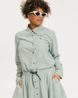 A woman wearing a green striped button-up shirt with a collar, sleeve cuffs, and side seam hem, paired with a matching skirt, sunglasses, and white shoes.