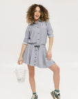 A woman wearing a long, button-up shirt with indigo stripes, featuring a collar, button-down front, and elastic toggles at the side seam hem and sleeve cuffs