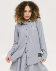 A woman wearing a long, button-up shirt with indigo stripes, featuring a collar, button-down front, and elastic toggles at the side seam hem and sleeve cuffs.