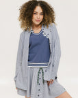 A woman wearing a long, button-up shirt with indigo stripes, featuring a collar, button-down front, and elastic toggles at the side seam hem and sleeve cuffs