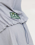 A woman wearing a long, button-up shirt with indigo stripes, featuring a collar, button-down front, and elastic toggles at the side seam hem and sleeve cuffs