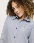 A woman wearing a long, button-up shirt with indigo stripes, featuring a collar, button-down front, and elastic toggles at the side seam hem and sleeve cuffs