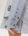 A woman wearing a long, button-up shirt with indigo stripes, featuring a collar, button-down front, and elastic toggles at the side seam hem and sleeve cuffs