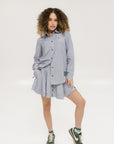 A woman wearing a long, button-up shirt with indigo stripes, featuring a collar, button-down front, and elastic toggles at the side seam hem and sleeve cuffs