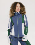 A female model wearing a blue and green color-blocked warm-up jacket with zipper pockets and a high neck collar.