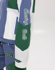A female model wearing a blue and green color-blocked warm-up jacket with zipper pockets and a high neck collar.