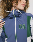 A female model wearing a blue and green color-blocked warm-up jacket with zipper pockets and a high neck collar.