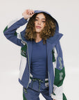 A female model wearing a blue and green color-blocked warm-up jacket with zipper pockets and a high neck collar.