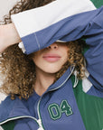 A female model wearing a blue and green color-blocked warm-up jacket with zipper pockets and a high neck collar.