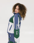 A female model wearing a blue and green color-blocked warm-up jacket with zipper pockets and a high neck collar.