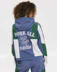 A female model wearing a blue and green color-blocked warm-up jacket with zipper pockets and a high neck collar.