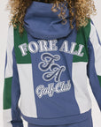 A female model wearing a blue and green color-blocked warm-up jacket with zipper pockets and a high neck collar.