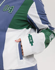 A female model wearing a blue and green color-blocked warm-up jacket with zipper pockets and a high neck collar.