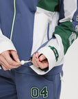 A female model wearing a blue and green color-blocked warm-up jacket with zipper pockets and a high neck collar.