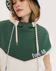 A person wearing a green and white sleeveless hoodie with a hood, matching green and white checkered hat, and green and white checkered skirt. The hoodie has white text and branding on it.