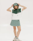 A person wearing a green and white sleeveless hoodie with a hood, matching green and white checkered hat, and green and white checkered skirt. The hoodie has white text and branding on it.