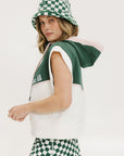 A person wearing a green and white sleeveless hoodie with a hood, matching green and white checkered hat, and green and white checkered skirt. The hoodie has white text and branding on it.