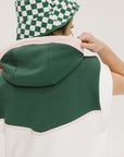 A person wearing a green sleeveless hoodie with a hood, paired with a checkered green and white hat.