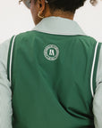 A woman wearing a green sleeveless windbreaker vest with a collar, featuring a white appliquéd logo on the left chest and silicone logos on the upper back.