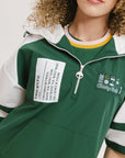 A woman wearing a green and white hoodie with a half-zip design, kangaroo pocket, and decorative woven ribbons.