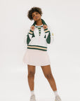 A woman wearing a white half zip sports top with green color blocking and ribbed detailing around the collar, cuffs, and hem. The top features half sleeve design with a signature logo on the chest.