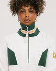 A woman wearing a white half zip sports top with green color blocking and ribbed detailing around the collar, cuffs, and hem. The top features half sleeve design with a signature logo on the chest.