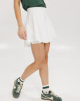 A person wearing a white skirt with a mesh back, paired with a green top and matching green and white sneakers. The skirt has a grippy elastic hem and built-in biddy shorties with pockets.