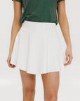 A person wearing a white skirt with a mesh back, paired with a green top and matching green and white sneakers. The skirt has a grippy elastic hem and built-in biddy shorties with pockets.