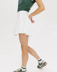 A person wearing a white skirt with a mesh back, paired with a green top and matching green and white sneakers. The skirt has a grippy elastic hem and built-in biddy shorties with pockets.