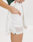 A person wearing a white skirt with a mesh back, paired with a green top and matching green and white sneakers. The skirt has a grippy elastic hem and built-in biddy shorties with pockets.