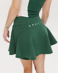 A woman is wearing a green mini skirt with a round drawstring waist and silicone logos. She is also wearing a pink top and white sneakers.