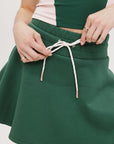 A woman is wearing a green mini skirt with a round drawstring waist and silicone logos. She is also wearing a pink top and white sneakers.
