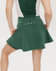 A woman is wearing a green mini skirt with a round drawstring waist and silicone logos. She is also wearing a pink top and white sneakers.