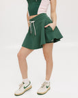 A woman is wearing a green mini skirt with a round drawstring waist and silicone logos. She is also wearing a pink top and white sneakers.