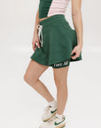 A woman is wearing a green mini skirt with a round drawstring waist and silicone logos. She is also wearing a pink top and white sneakers.