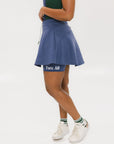 A woman is wearing a navy blue skirt with a built-in pair of biddy shorties featuring a white and green logo. She has a round drawstring cord at the waist and stitched logos on the back.
