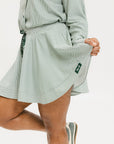 Model wearing the patty skirt in green seersucker. Featuring a green drawstring, buttons down the front, and a fore all logo on the bottom of the skirt.