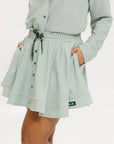 Model wearing the patty skirt in green seersucker. Featuring a green drawstring, buttons down the front, and a fore all logo on the bottom of the skirt.