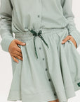 Model wearing the patty skirt in green seersucker. Featuring a green drawstring, buttons down the front, and a fore all logo on the bottom of the skirt.