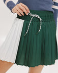 A woman wearing a pleated green and white skirt with a round drawstring waist, colorblocking, and a 'Chasing Balls' icon patch on the back.