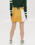 A person standing against a white background wearing a rose pleated skirt in mustard color paired with a green sweater with white stripes, and white sneakers.