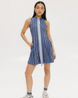 A woman is wearing a sleeveless, pleated, pinstripe indigo dress with a halter neck, side seam pockets, and a full-length two-way zipper at the center front. She has paired the dress with white sneakers and is standing against a white background.