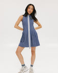 A woman is wearing a sleeveless, pleated, pinstripe indigo dress with a halter neck, side seam pockets, and a full-length two-way zipper at the center front. She has paired the dress with white sneakers and is standing against a white background.