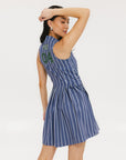 A woman is wearing a sleeveless, pleated, pinstripe indigo dress with a halter neck, side seam pockets, and a full-length two-way zipper at the center front. She has paired the dress with white sneakers and is standing against a white background.