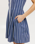 A woman is wearing a sleeveless, pleated, pinstripe indigo dress with a halter neck, side seam pockets, and a full-length two-way zipper at the center front. She has paired the dress with white sneakers and is standing against a white background.