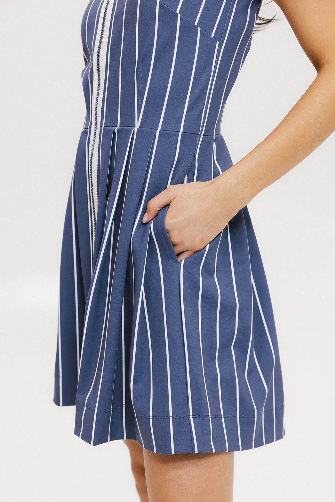 A woman is wearing a sleeveless, pleated, pinstripe indigo dress with a halter neck, side seam pockets, and a full-length two-way zipper at the center front. She has paired the dress with white sneakers and is standing against a white background.